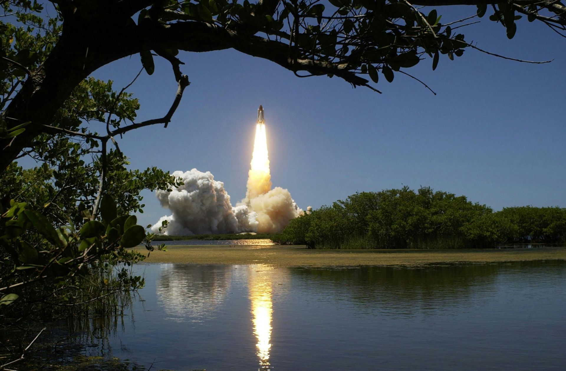 A dramatic rocket launch with smoke and flames, reflected in calm waters and framed by trees.